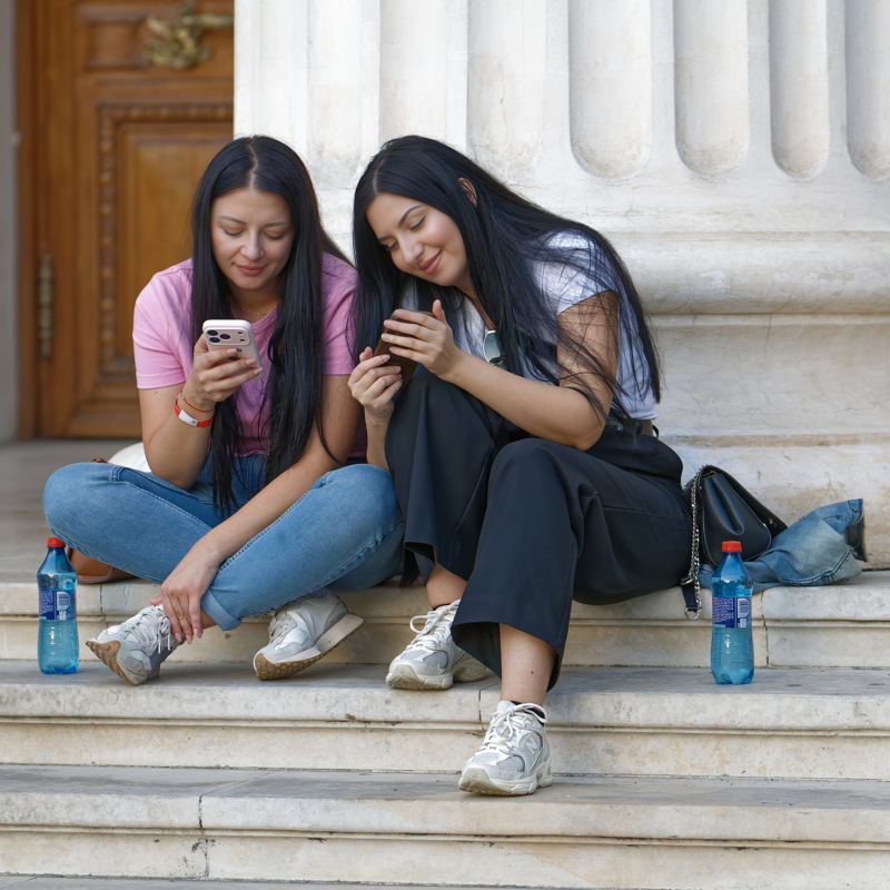 27 - Two women with long dark hair sit casually on stone steps while looking at their mobile phones